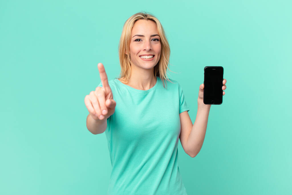 jovem mulher loira sorrindo orgulhosamente e confiantemente fazendo número um e segurando um telefone inteligente - Foto, Imagem