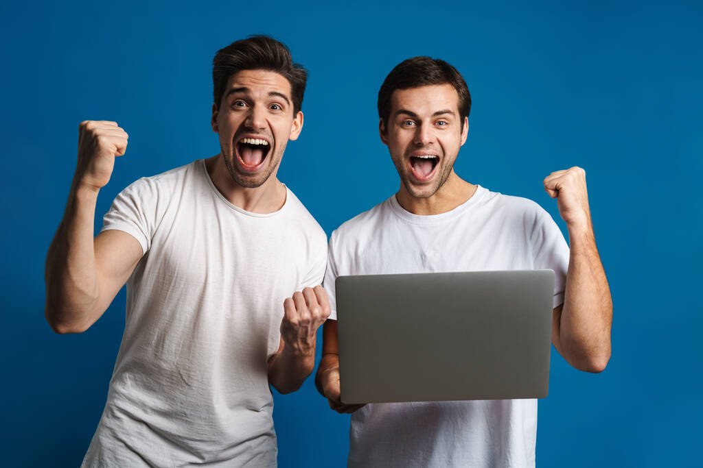 Aufgeregt machen zwei Jungs Siegergeste, während sie mit Laptop isoliert vor blauem Hintergrund posieren - Foto, Bild