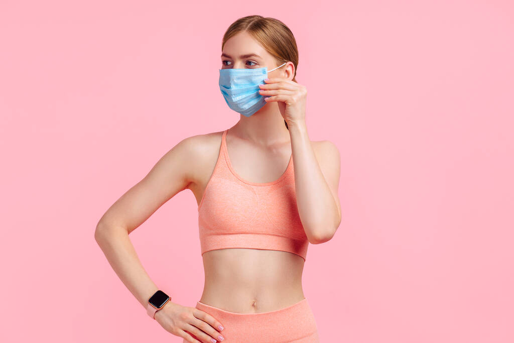 Beautiful young fitness woman wears protective mask to prevent corona virus, sportswoman with perfect body wears sportswear for workouts, on isolated pink background - Photo, Image