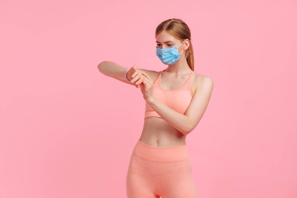 Jeune femme sportive portant un masque protecteur contre la maladie du coronavirus, utilisant une montre connectée, vérifiant l'activité d'un bracelet, sur un fond rose - Photo, image