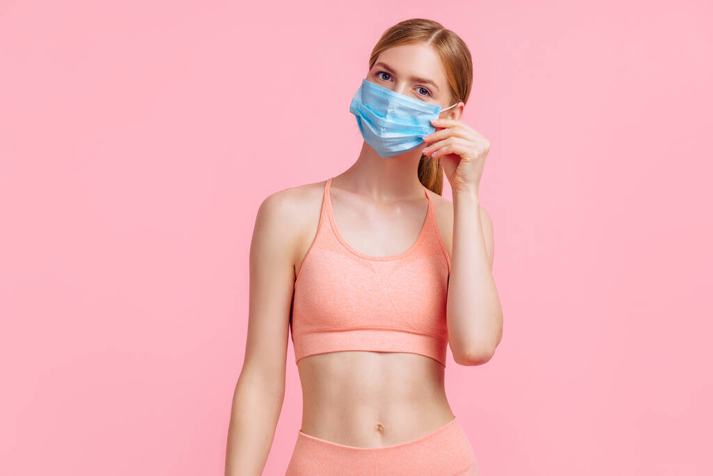 Aantrekkelijke fitnessvrouw met beschermend masker van het COVID-virus, lachende vrouw na fitnesstraining, op roze achtergrond, concept van gezonde levensstijl - Foto, afbeelding