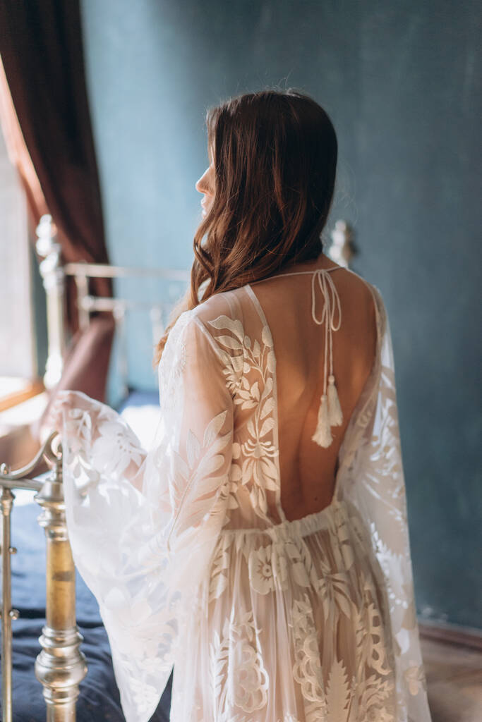 chica en un hermoso vestido floral sensual bohemio blanco de pie a media vuelta junto a la cama en luces y sombras con su espalda a la cámara - Foto, Imagen