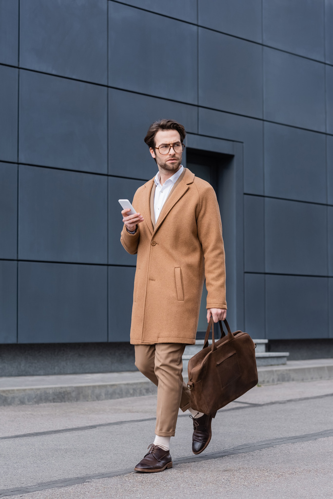 young man in glasses walking with briefcase and smartphone near building  - Fotografie, Obrázek