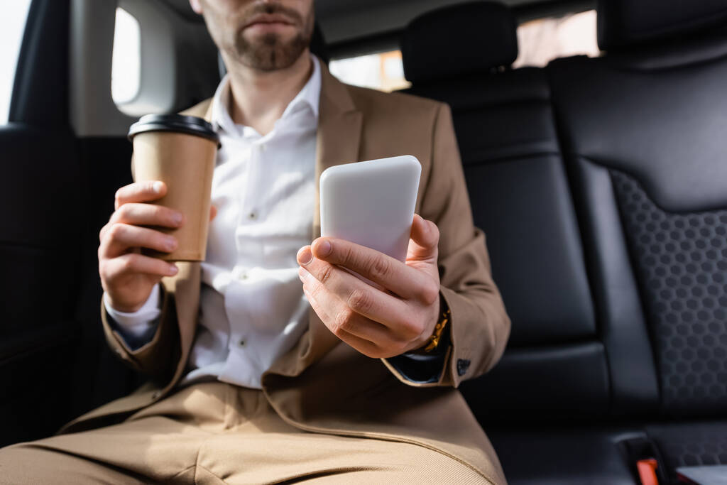 cropped view of man in suit holding paper cup and smartphone in car  - Fotoğraf, Görsel