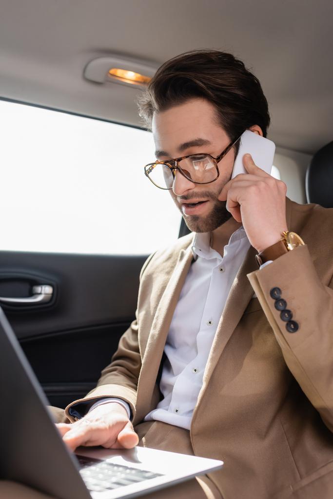 businessman working remotely with laptop while talking on smartphone in car - Foto, afbeelding