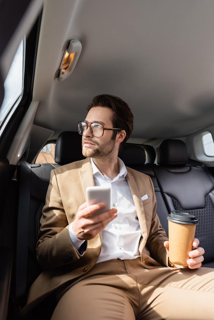 uomo d'affari pensieroso in tuta e occhiali con tazza di carta e smartphone in auto  - Foto, immagini