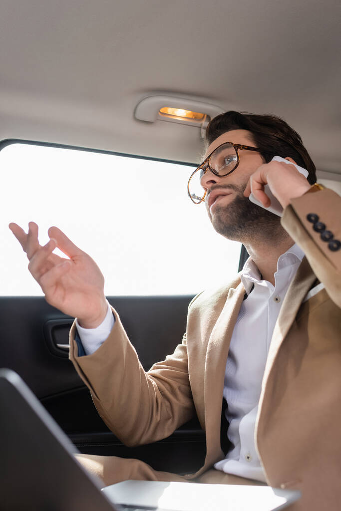 vista de ángulo bajo de hombre de negocios en gafas hablando en el teléfono inteligente cerca de ordenador portátil borroso en el coche - Foto, imagen