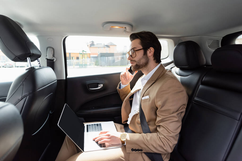 車の中で携帯電話で話をしながらノートパソコンでリモートで作業する眼鏡のビジネスマン - 写真・画像