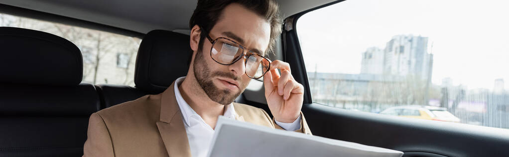 uomo sicuro di sé che regola gli occhiali e legge il giornale aziendale in auto, banner - Foto, immagini