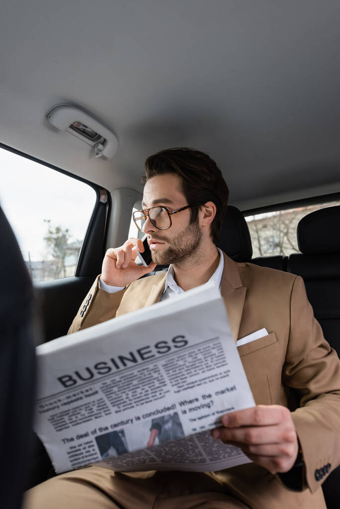 homem confiante em óculos falando no smartphone e segurando jornal de negócios no carro  - Foto, Imagem