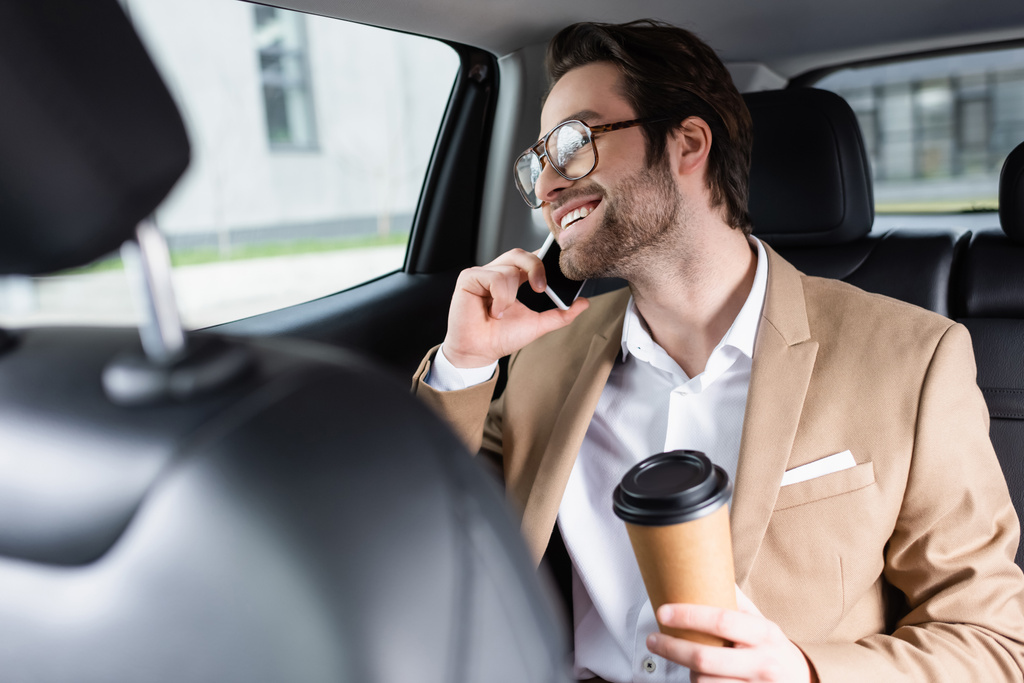 joyful man in suit and glasses holding paper cup and talking on smartphone in car  - Zdjęcie, obraz