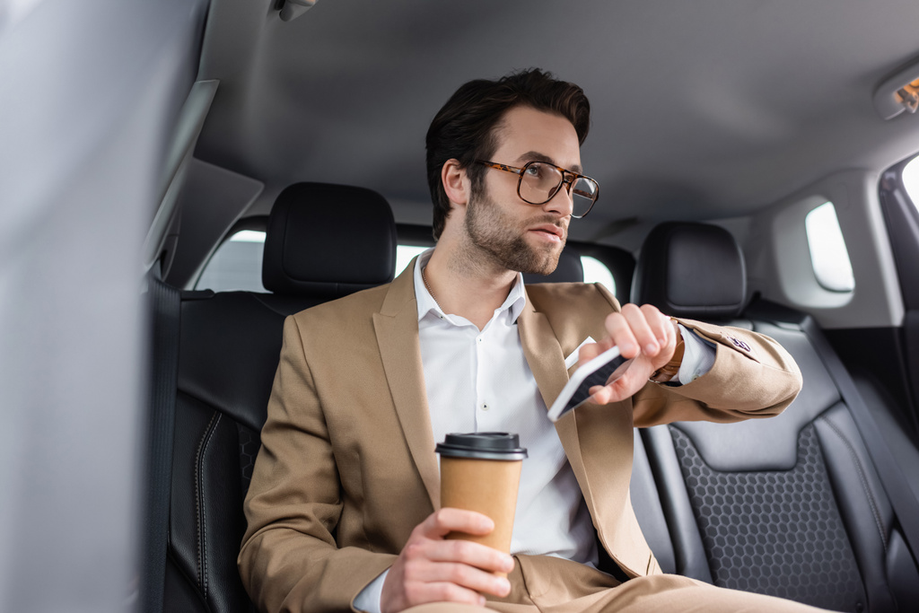 businessman in beige suit and glasses holding paper cup and smartphone in car  - Foto, afbeelding