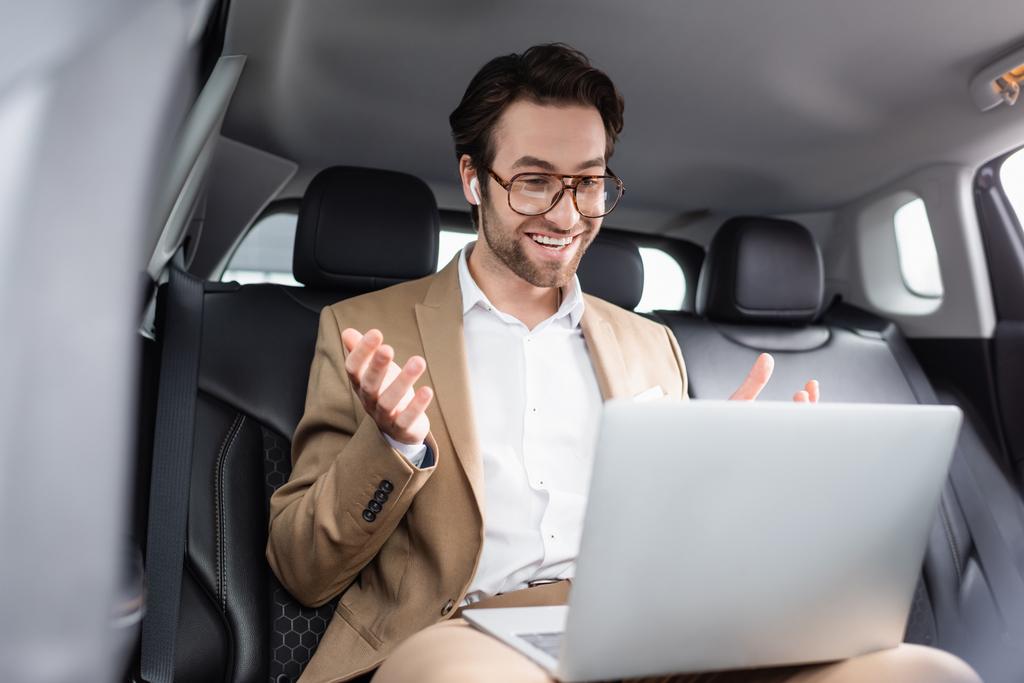 улыбающийся бизнесмен в очках и беспроводных наушниках жестикулирует во время видеозвонка в машине  - Фото, изображение