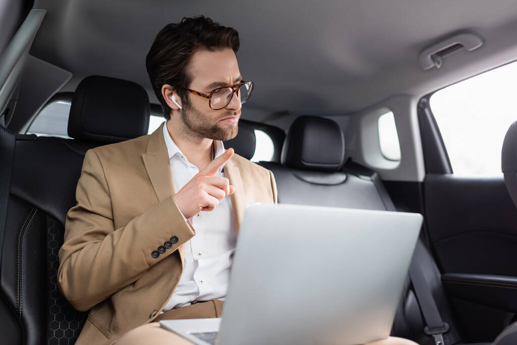 Geschäftsmann mit Brille und drahtlosen Kopfhörern zeigt bei Videoanruf im Auto mit dem Finger  - Foto, Bild