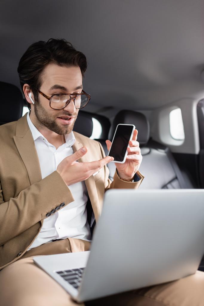 Geschäftsmann mit Brille und drahtlosem Kopfhörer zeigt bei Videoanruf im Auto auf Smartphone  - Foto, Bild