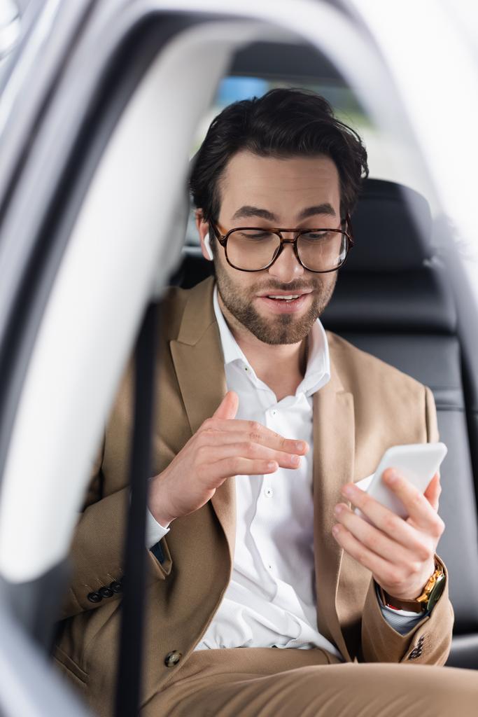 businessman in glasses and wireless earphones gesturing during video call on smartphone inside of car  - Photo, Image