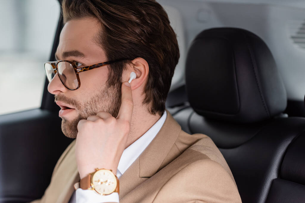 bearded man in glasses and beige jacket adjusting wireless earphones in car - Foto, afbeelding