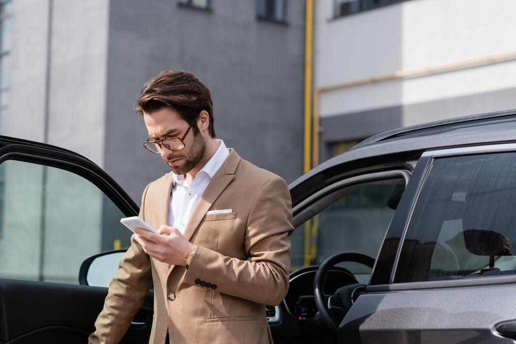 bearded man in suit and glasses looking at cellphone and standing near car with opened door - Foto, afbeelding