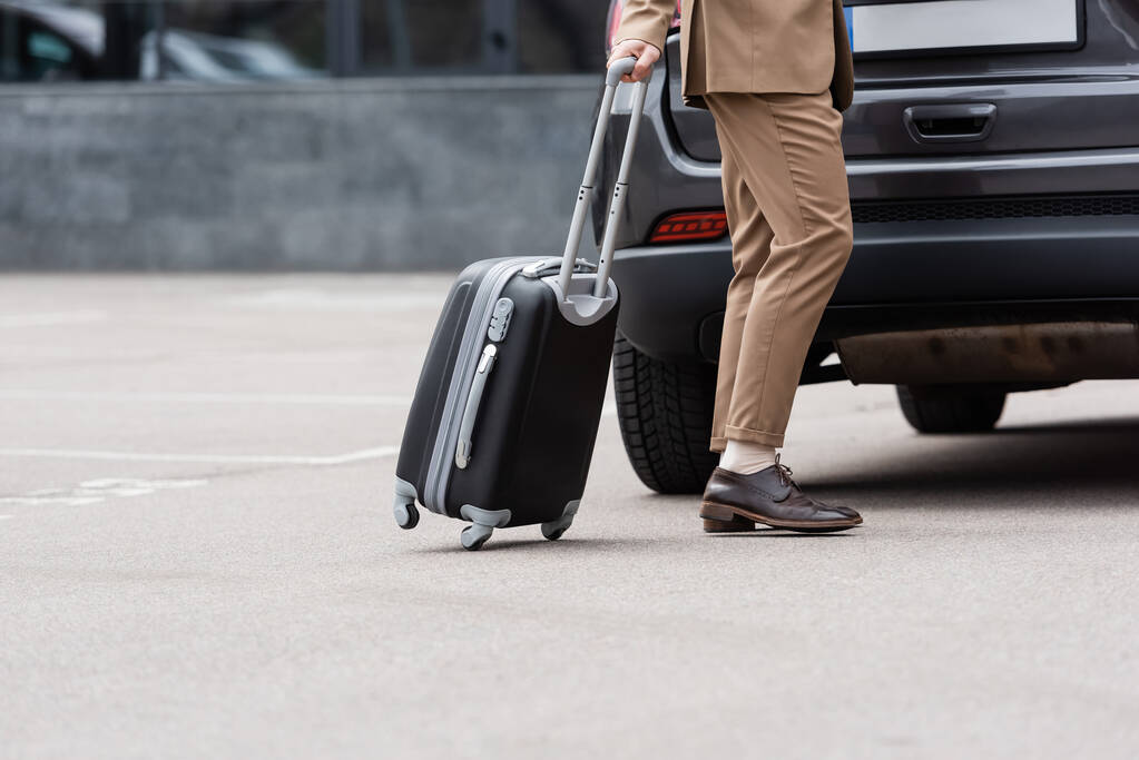 vista recortada del hombre en traje caminando cerca del coche con la maleta  - Foto, Imagen