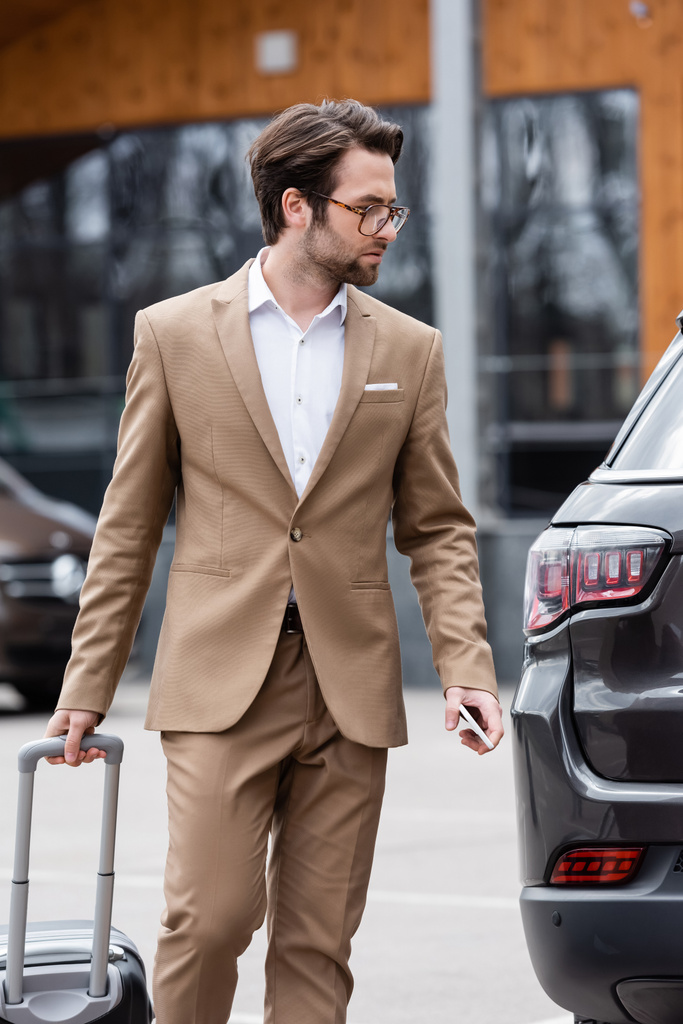 férfi bézs öltöny és szemüveg gazdaság smartphone és az autó közelében áll a bőrönd  - Fotó, kép