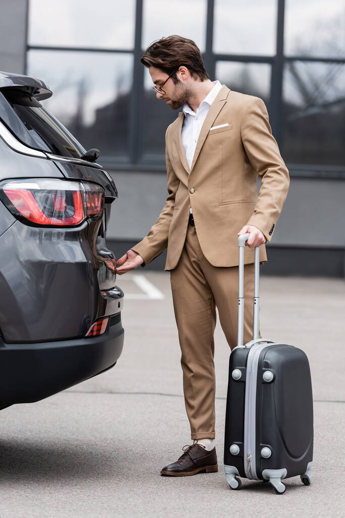 человек в костюме стоит с чемоданом и открывает багажник автомобиля  - Фото, изображение