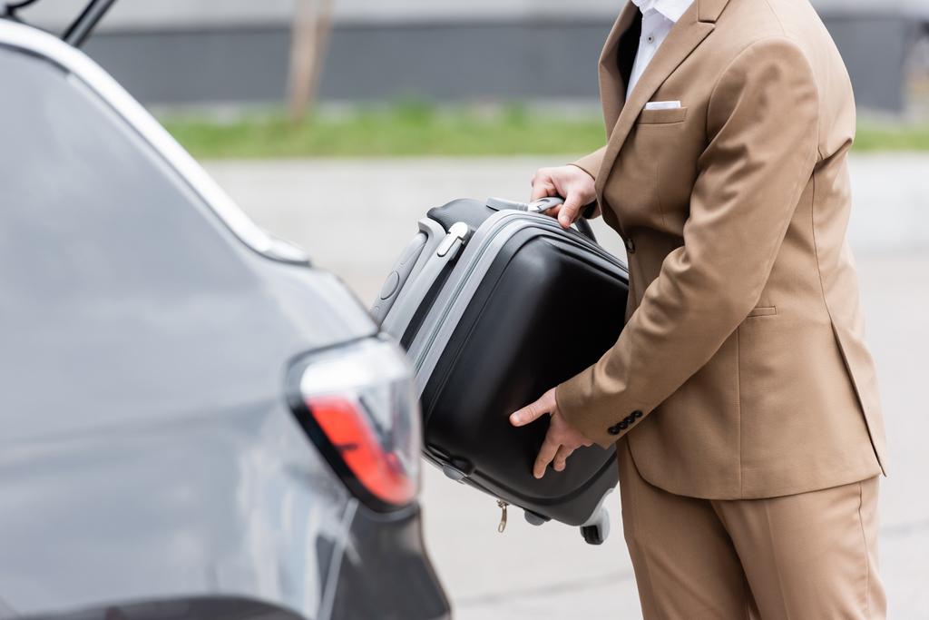 cropped view of man in suit putting suitcase in car trunk  - Foto, afbeelding
