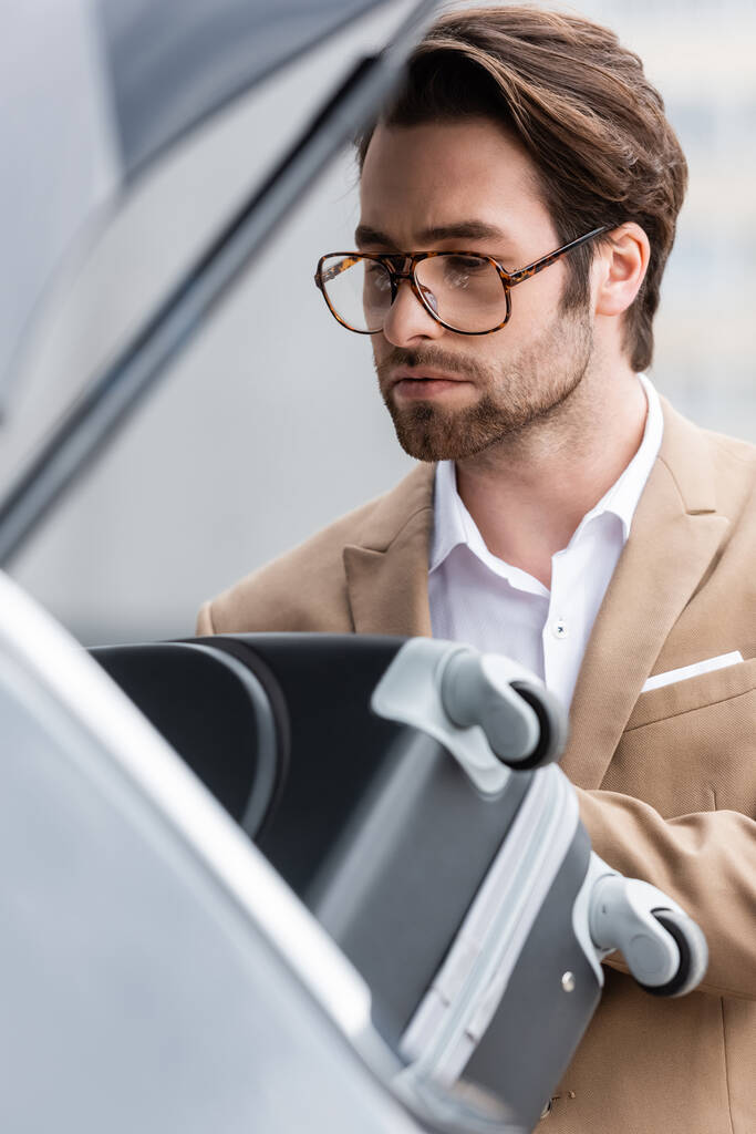бородатый мужчина в очках и костюме кладет чемодан в багажник размытой машины  - Фото, изображение