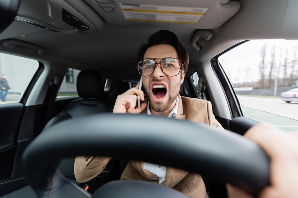 homem zangado falando no smartphone enquanto dirige carro em primeiro plano desfocado  - Foto, Imagem