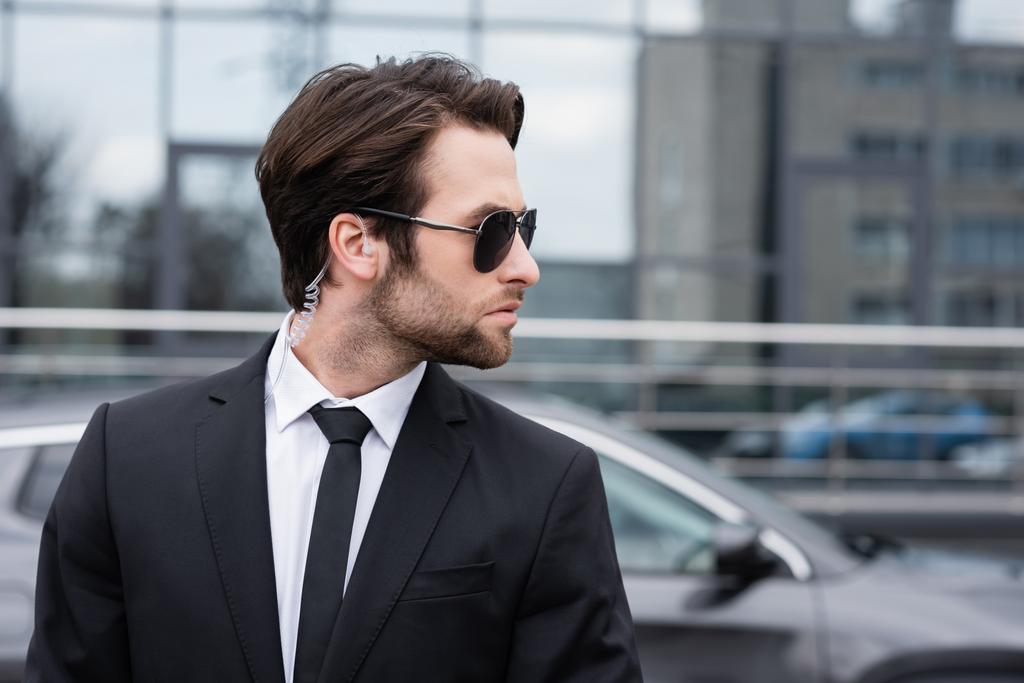 Seitenansicht des bärtigen Leibwächters in Anzug und Sonnenbrille mit Sicherheitsbügel in der Nähe moderner Autos  - Foto, Bild