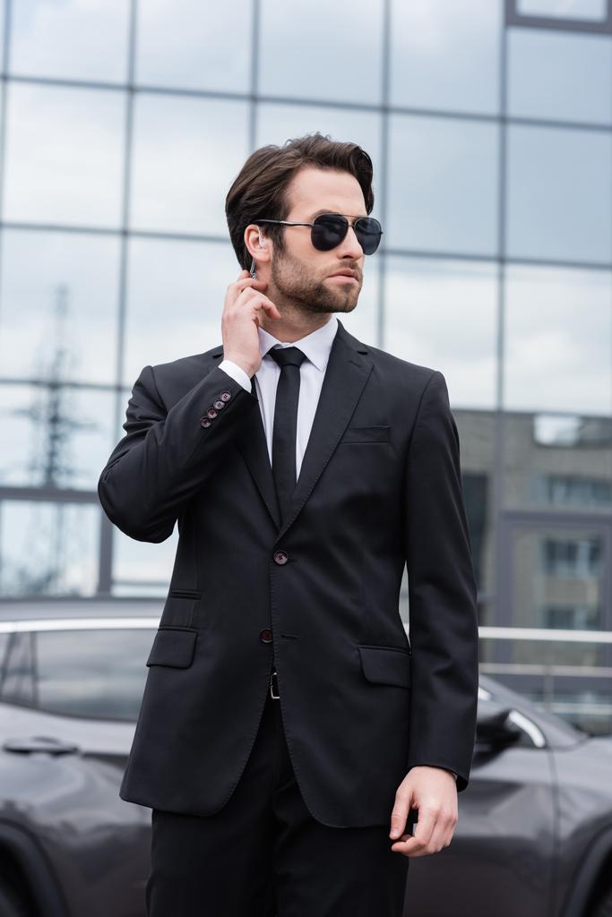 bearded bodyguard in suit and sunglasses with security earpiece near modern building on blurred background  - Photo, Image
