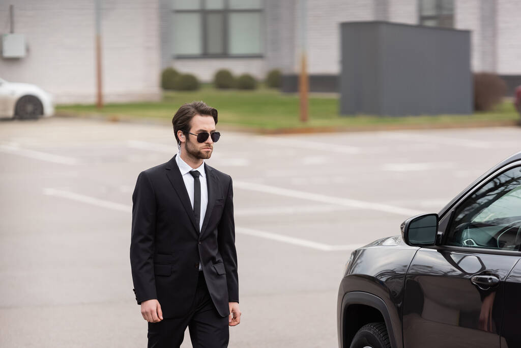 近代的な自動車の近くを歩くセキュリティイヤーピース付きのスーツとサングラスの髭ボディガード  - 写真・画像