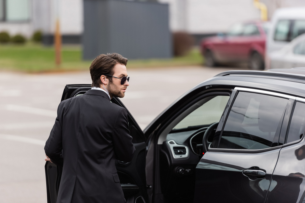 bearded bodyguard in suit and sunglasses with security earpiece near opened door of modern auto  - Foto, afbeelding