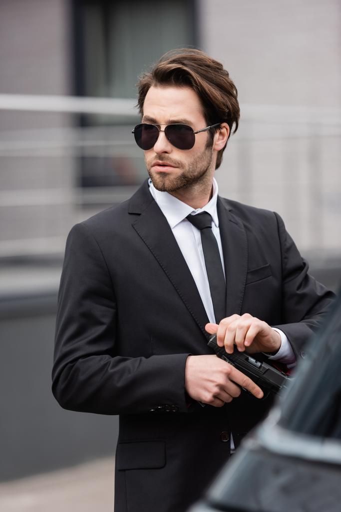 bearded bodyguard in sunglasses holding gun near blurred auto  - Fotoğraf, Görsel