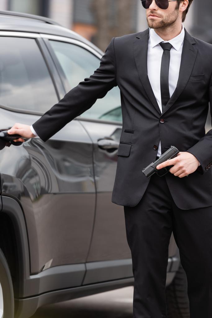 Ausgeschnittene Sicht auf Schutzmann in Anzug und Sonnenbrille mit Pistole in der Nähe verschwommenes modernes Auto - Foto, Bild