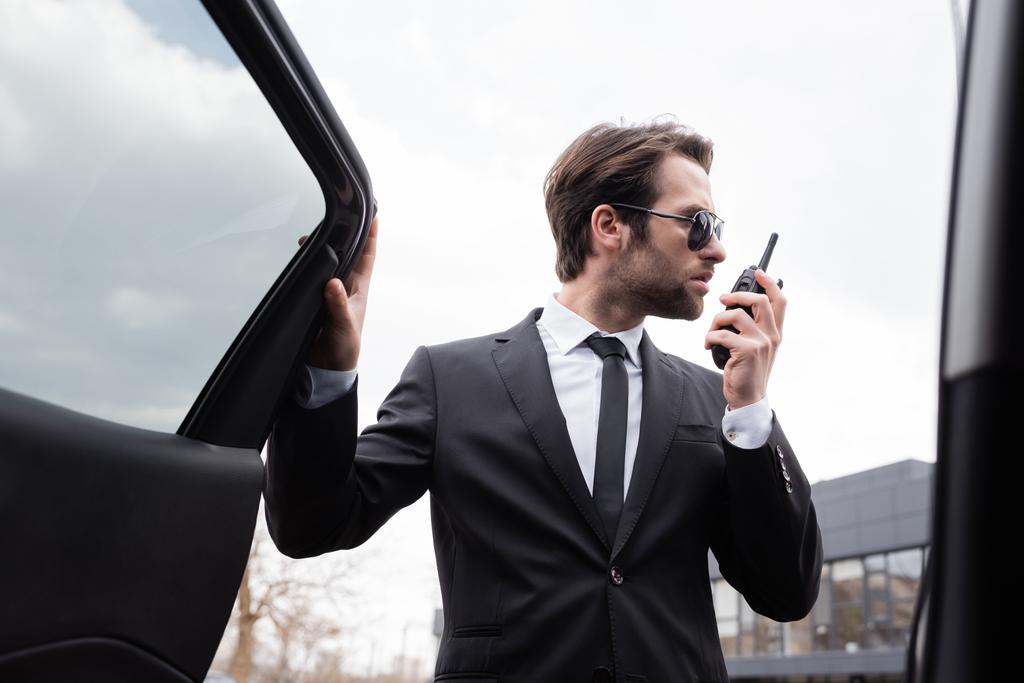 vista lateral de guardaespaldas en traje y gafas de sol usando walkie talkie cerca de coche moderno - Foto, Imagen