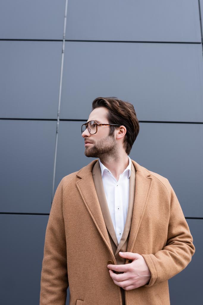 parrakas liikemies lasit ja beige takki seisoo rakennuksen ulkopuolella - Valokuva, kuva