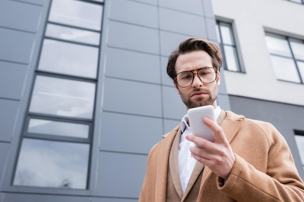 vista de ángulo bajo del hombre de negocios barbudo en gafas y abrigo beige usando teléfono celular cerca del edificio exterior  - Foto, imagen