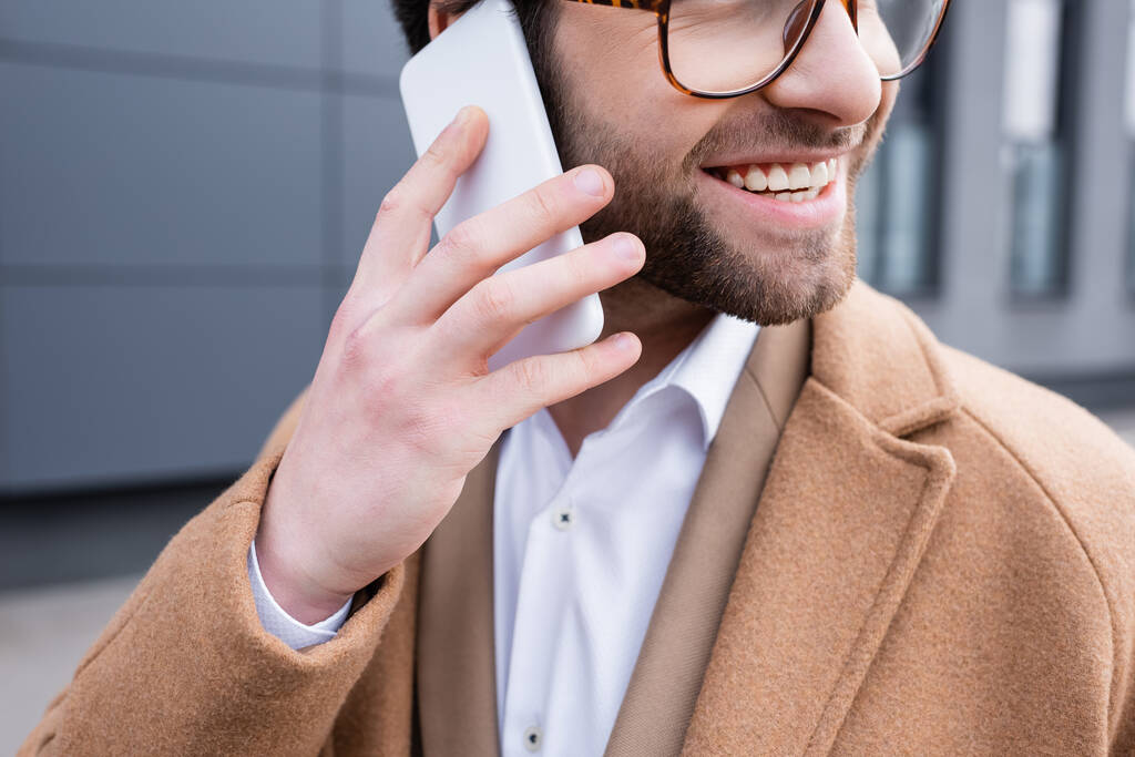 rajattu näkymä onnellinen liikemies lasit ja beige takki puhuu kännykkään lähellä rakennuksen ulkopuolella  - Valokuva, kuva