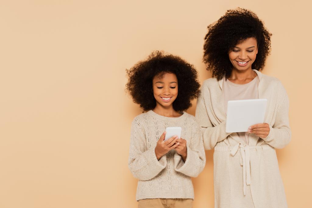 smiling african american preteen daughter and adult mother holding cellphone and tablet in hands isolated on beige - Photo, Image
