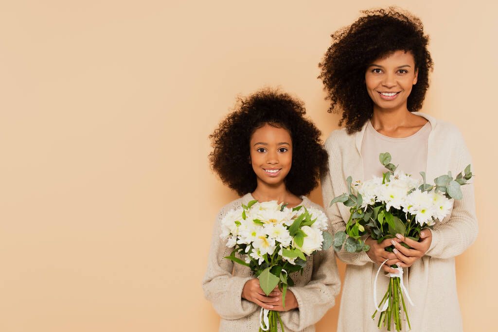 hija preadolescente afroamericana y madre adulta sosteniendo ramos de margaritas aisladas en beige - Foto, Imagen