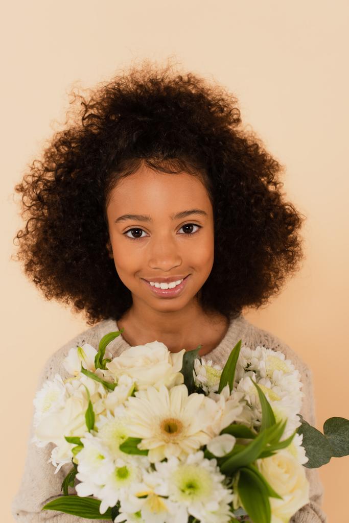 retrato de niña preadolescente afroamericana sonriente con ramo de flores en manos aisladas en beige - Foto, Imagen