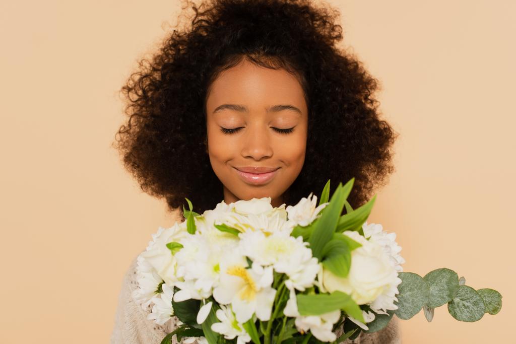 happy preteen african american girl smelling bouquet of daisies with closed eyes isolated on beige - Photo, Image