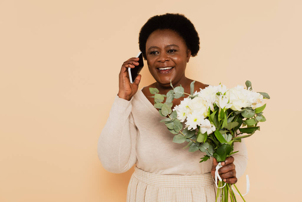 Afrikaans amerikaanse dame van middelbare leeftijd spreken op smartphone in en het houden van boeket van bloemen in de hand op beige achtergrond - Foto, afbeelding
