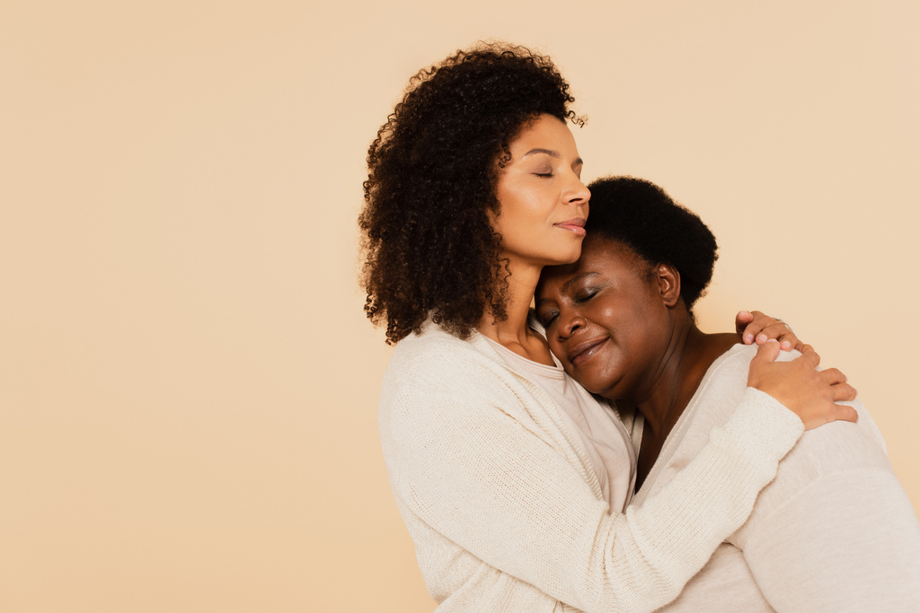 Αφροαμερικανή ενήλικη κόρη που αγκαλιάζει μεσήλικη μητέρα με κλειστά μάτια απομονωμένη στο μπεζ - Φωτογραφία, εικόνα