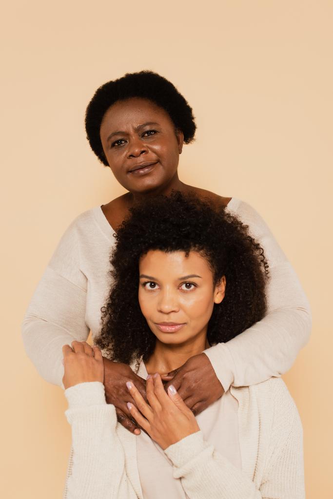 portrait of african american middle aged mother and adult daughter hugging and holding hands isolated on beige - Photo, Image