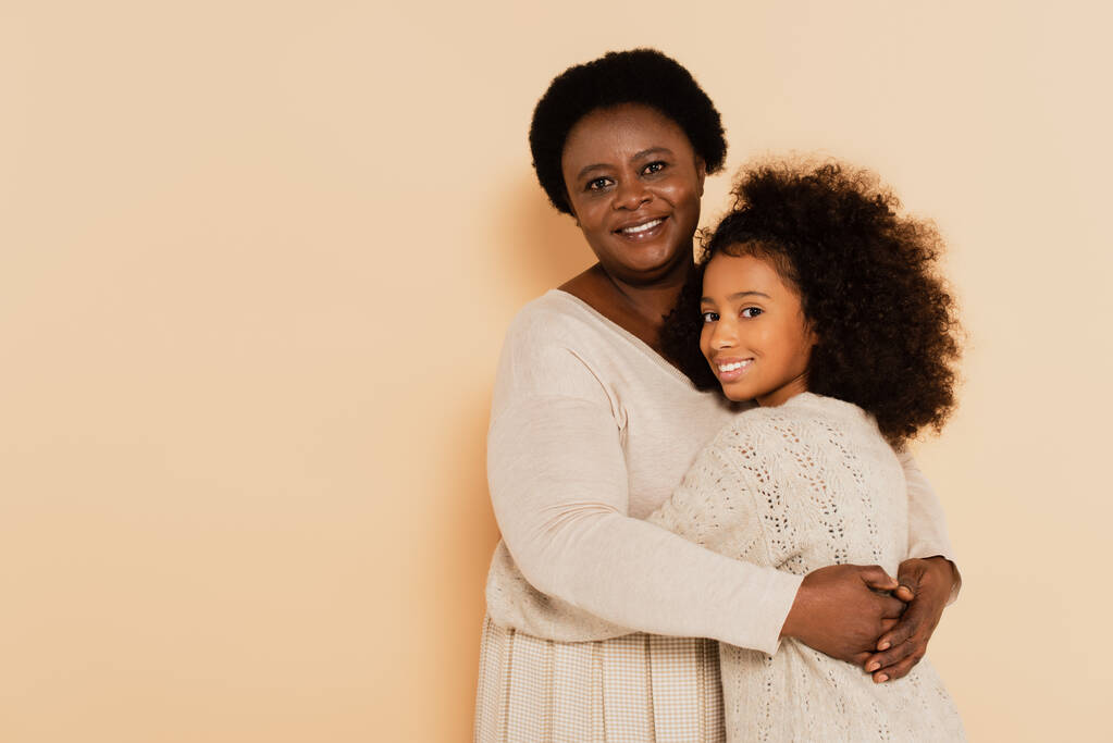 smiling african american grandmother hugging granddaughter on beige background - Photo, Image