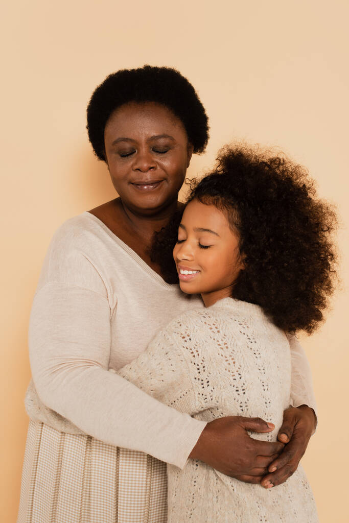 Afrikaans amerikaans grootmoeder knuffelen kleindochter met gesloten ogen op beige achtergrond - Foto, afbeelding