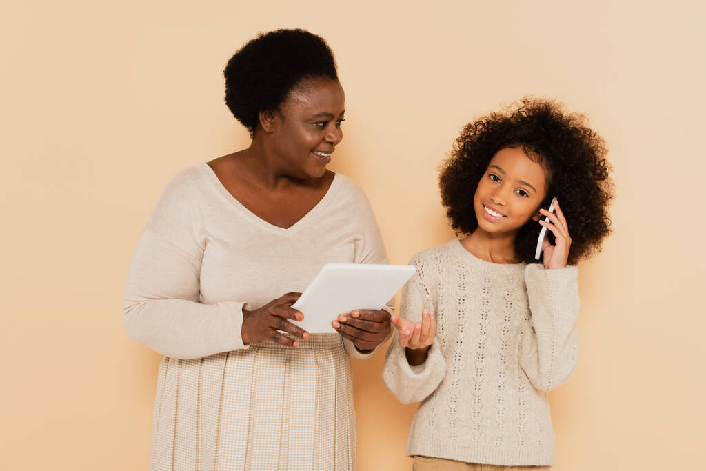 afrikkalainen amerikkalainen tyttärentytär puhuu kännykällä lähellä isoäitiä tabletti käsissä beige tausta - Valokuva, kuva
