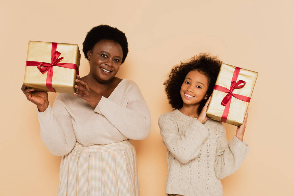 щаслива афроамериканська онука і бабуся тримає подарункові коробки біля голів на бежевому фоні
 - Фото, зображення