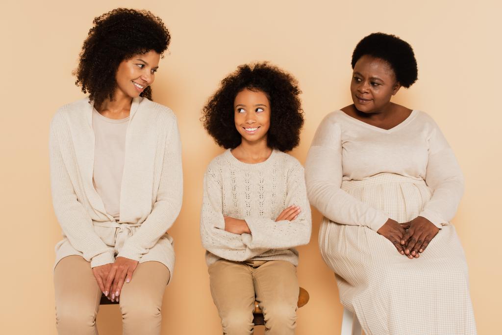 afro-américaine petite-fille assise avec les bras croisés entre grand-mère et mère sur fond beige - Photo, image